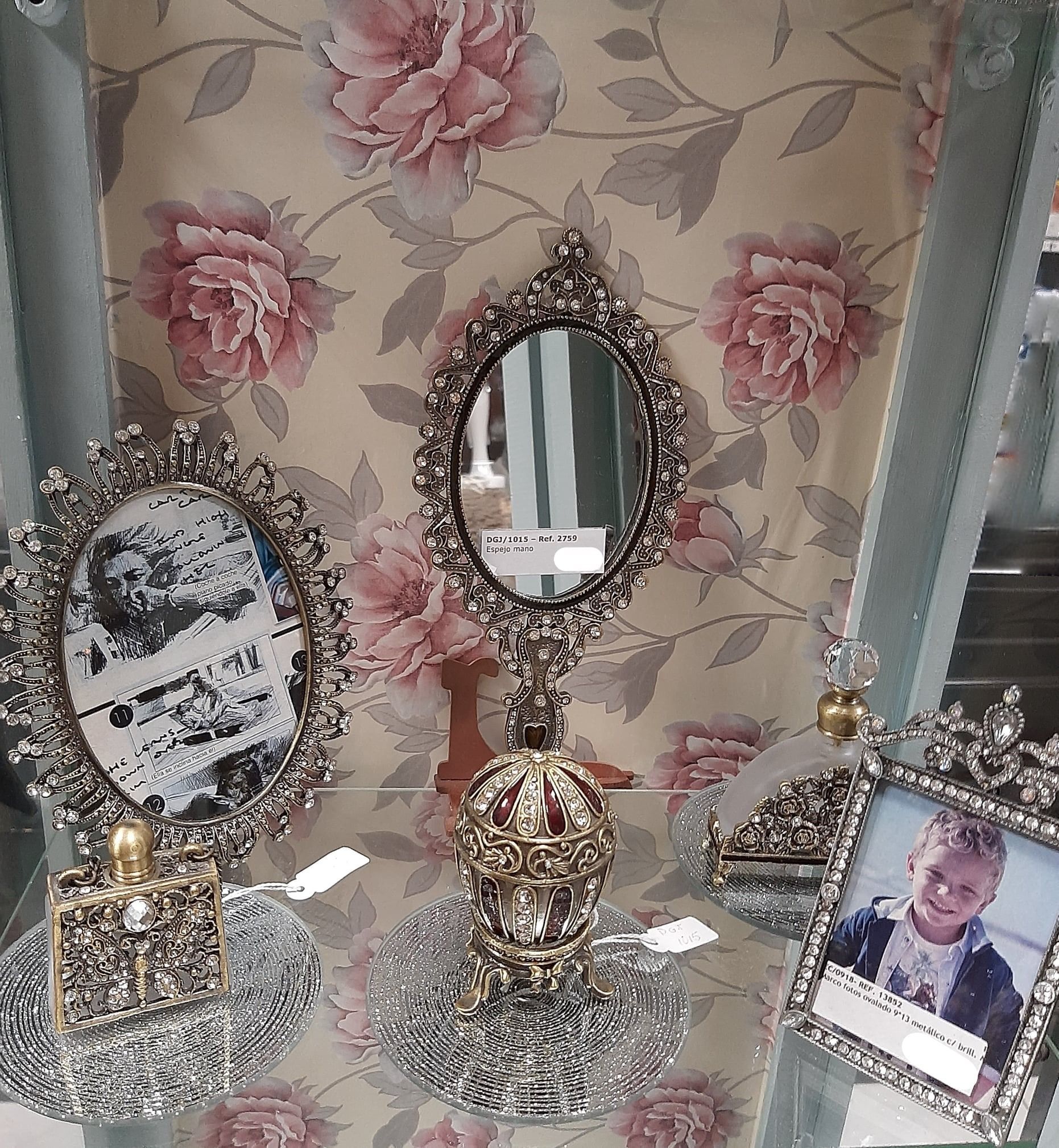 Exhibición de espejos adornados con marcos de plata y frascos de perfume en un estante de vidrio, con fondo de papel tapiz floral.