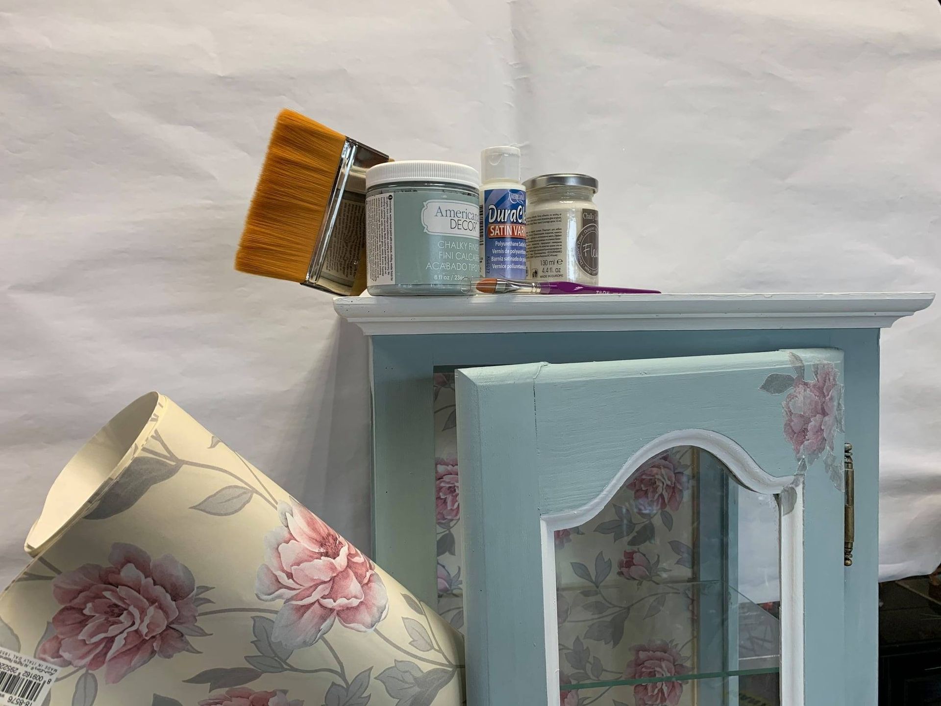 Un gabinete azul con decoración floral, suministros de pintura y una lámpara sobre un fondo blanco.