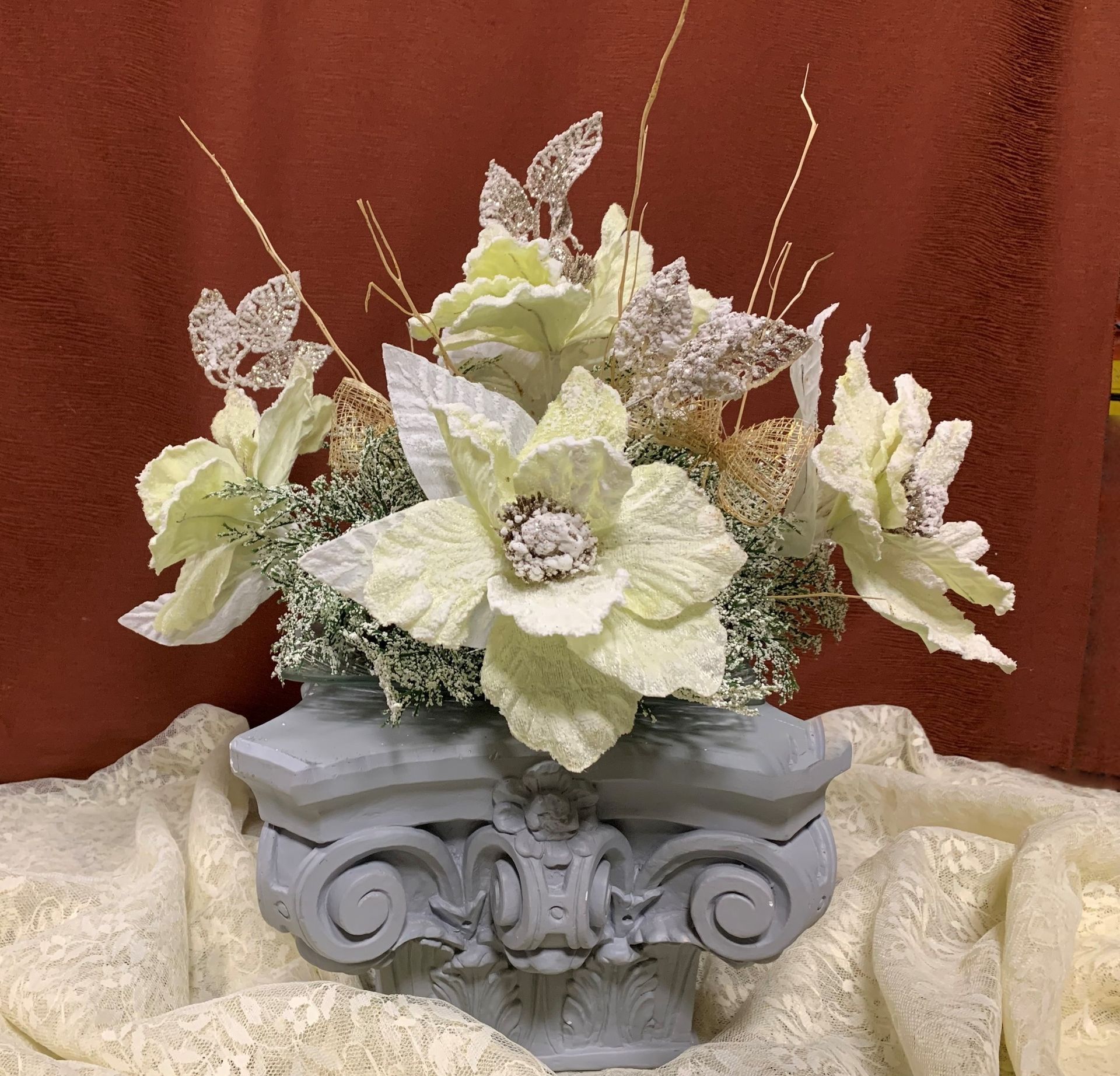 Arreglo floral en pedestal gris, con flores de color verde claro y blanco y ramas decorativas sobre un fondo marrón.