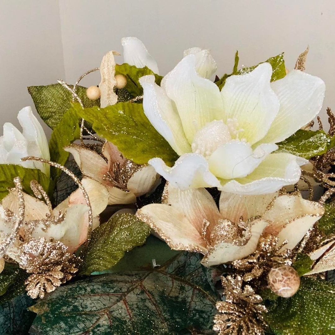 Primer plano de flores artificiales blancas y doradas con hojas verdes y brillo dorado sobre una base decorativa.