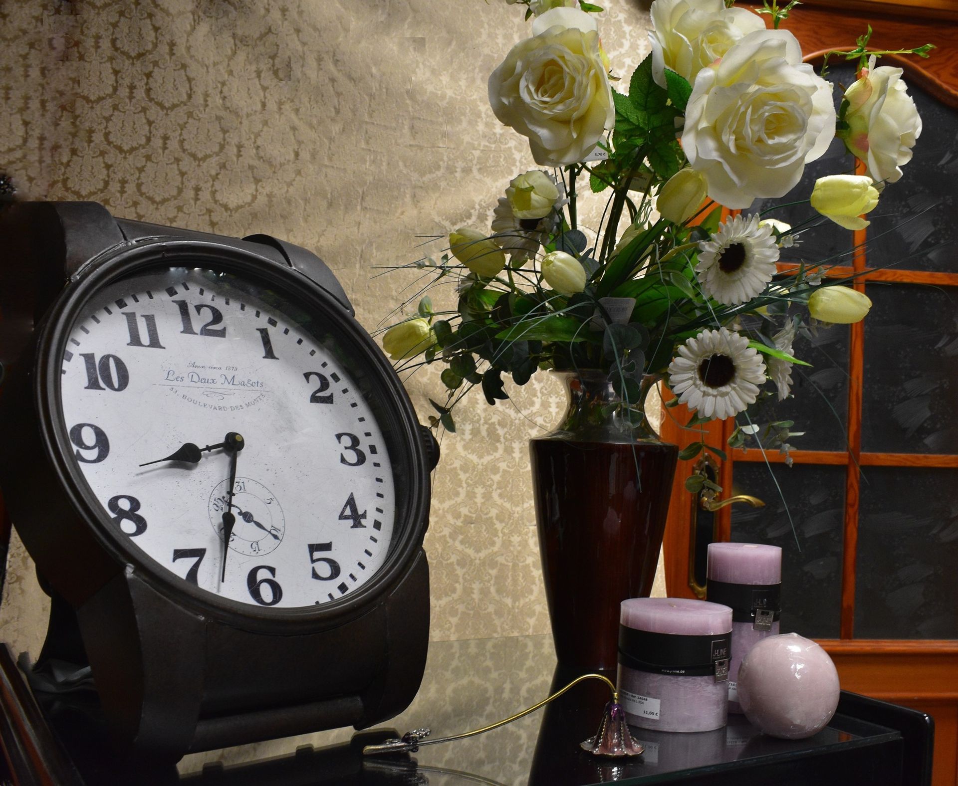 Reloj negro, esfera blanca, flores en jarrón, velas en la mesa.