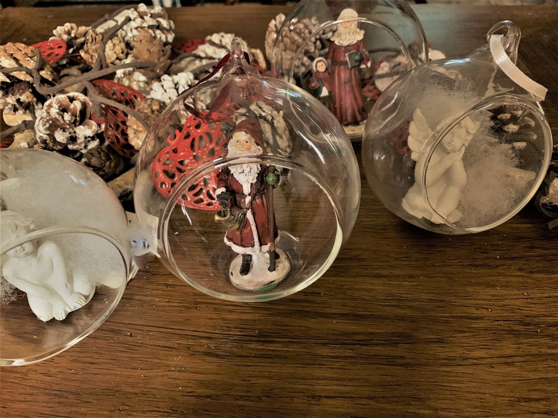 Adornos navideños: Figuras de Papá Noel, ángel y mujer dentro de globos de cristal transparente, sobre madera con piñas.