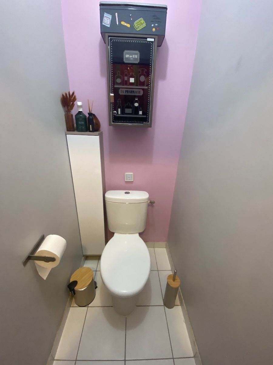 Une petite salle de bains avec des toilettes blanches, un porte-papier toilette, une armoire et un distributeur fixé sur un mur rose pâle.