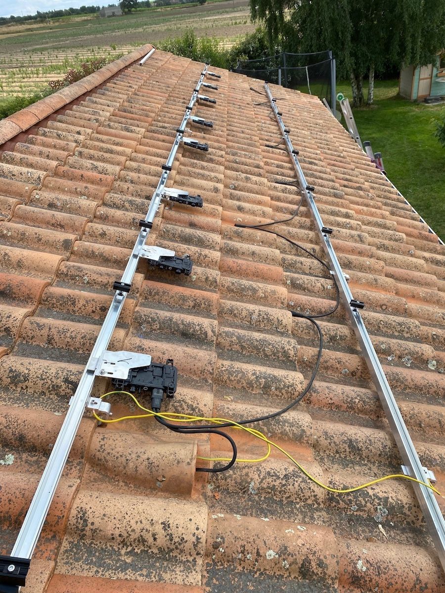 Deux rails métalliques parallèles pour le montage de panneaux solaires sont installés sur un toit en tuiles de terre cuite, laissant apparaître le câblage électrique.