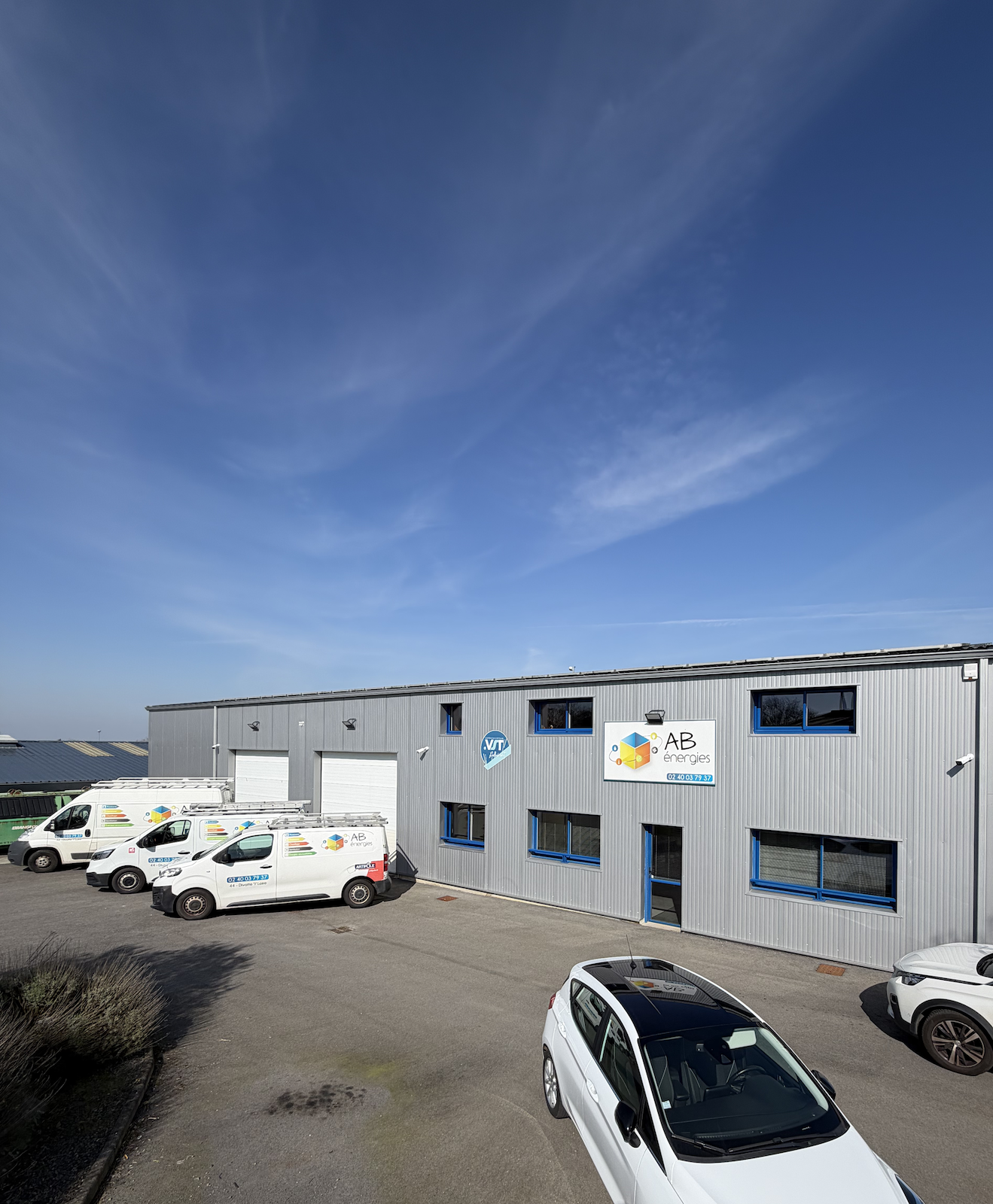 Un bâtiment industriel gris avec des fourgonnettes d'entreprise et une voiture garées, le tout sous un ciel bleu azur.