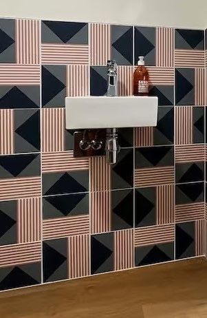 Un petit lavabo blanc mural avec robinet et flacon de savon, adossé à un mur carrelé à motifs géométriques.