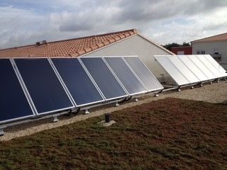 Des rangées de panneaux solaires thermiques de couleurs claires et foncées sont installées sur un toit plat recouvert de gravier, à côté d'une maison.