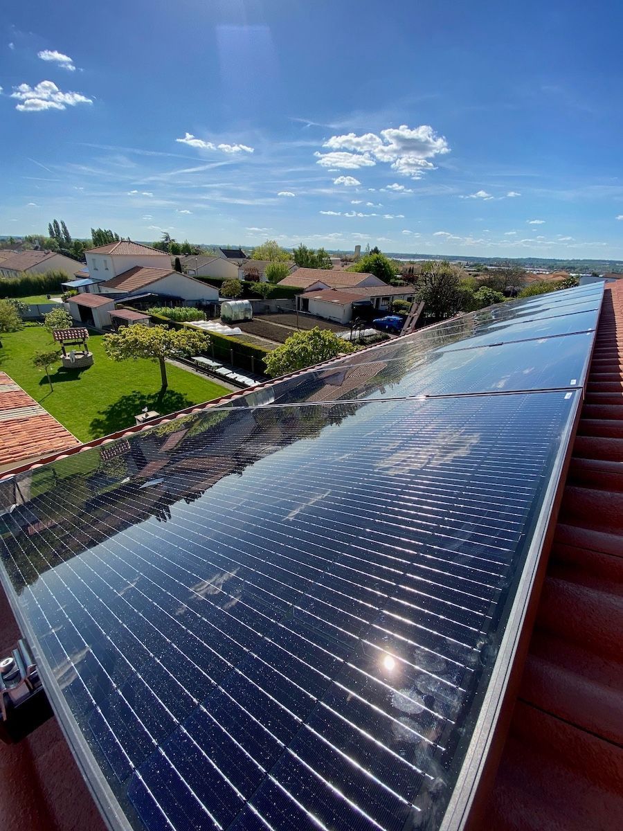 Des panneaux solaires sur un toit de tuiles sous un ciel d'un bleu éclatant, donnant sur une cour arrière résidentielle avec des arbres et des maisons.