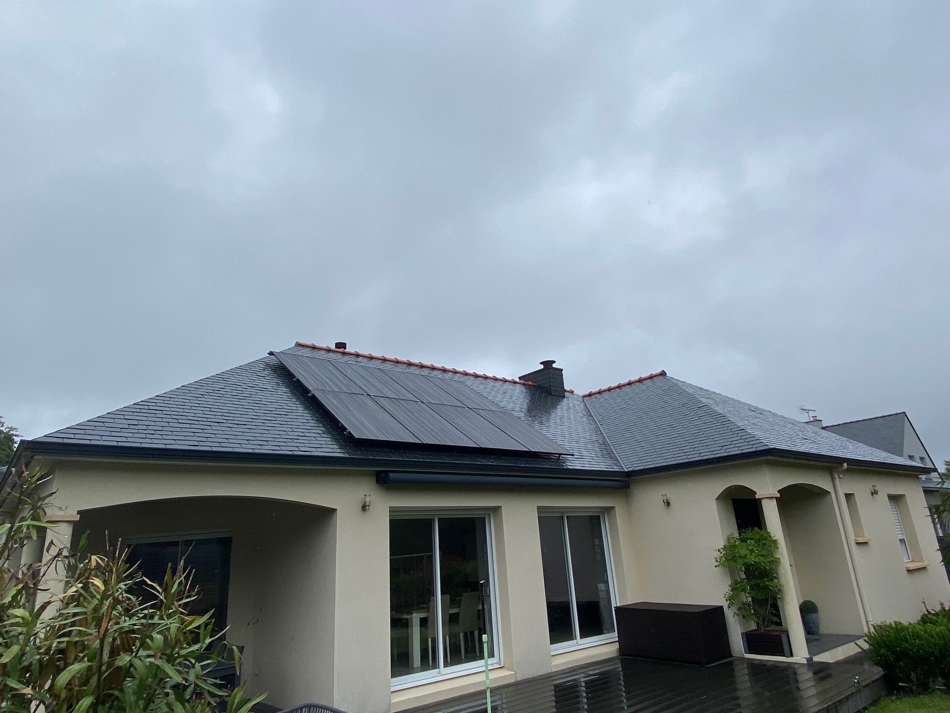 Une maison beige de plain-pied avec un toit de tuiles sombres et une partie recouverte de panneaux solaires, sous un ciel nuageux et couvert.