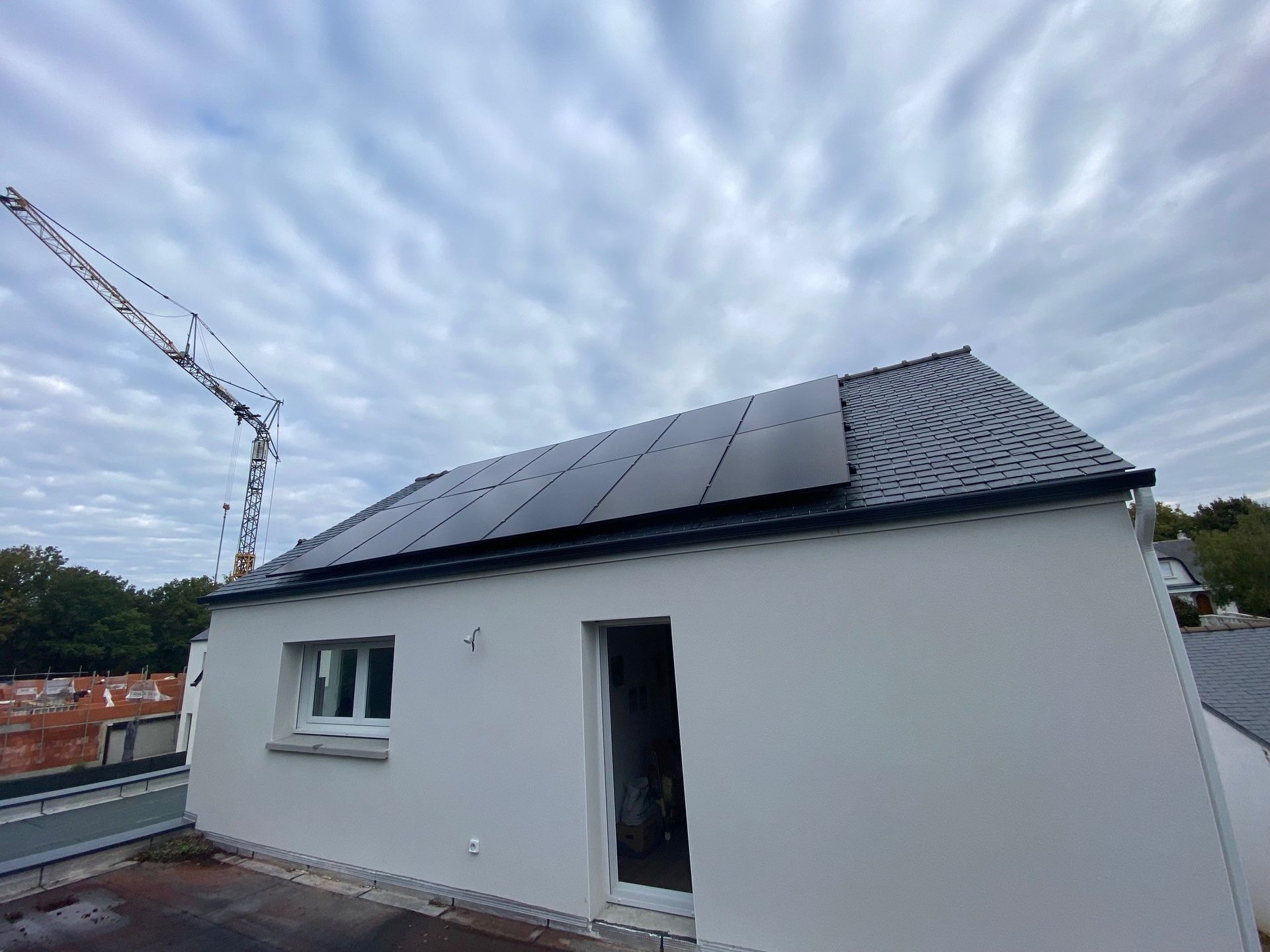 Extérieur d'une maison blanche moderne avec un toit équipé de panneaux solaires noirs, sous un ciel nuageux avec une grue au loin.