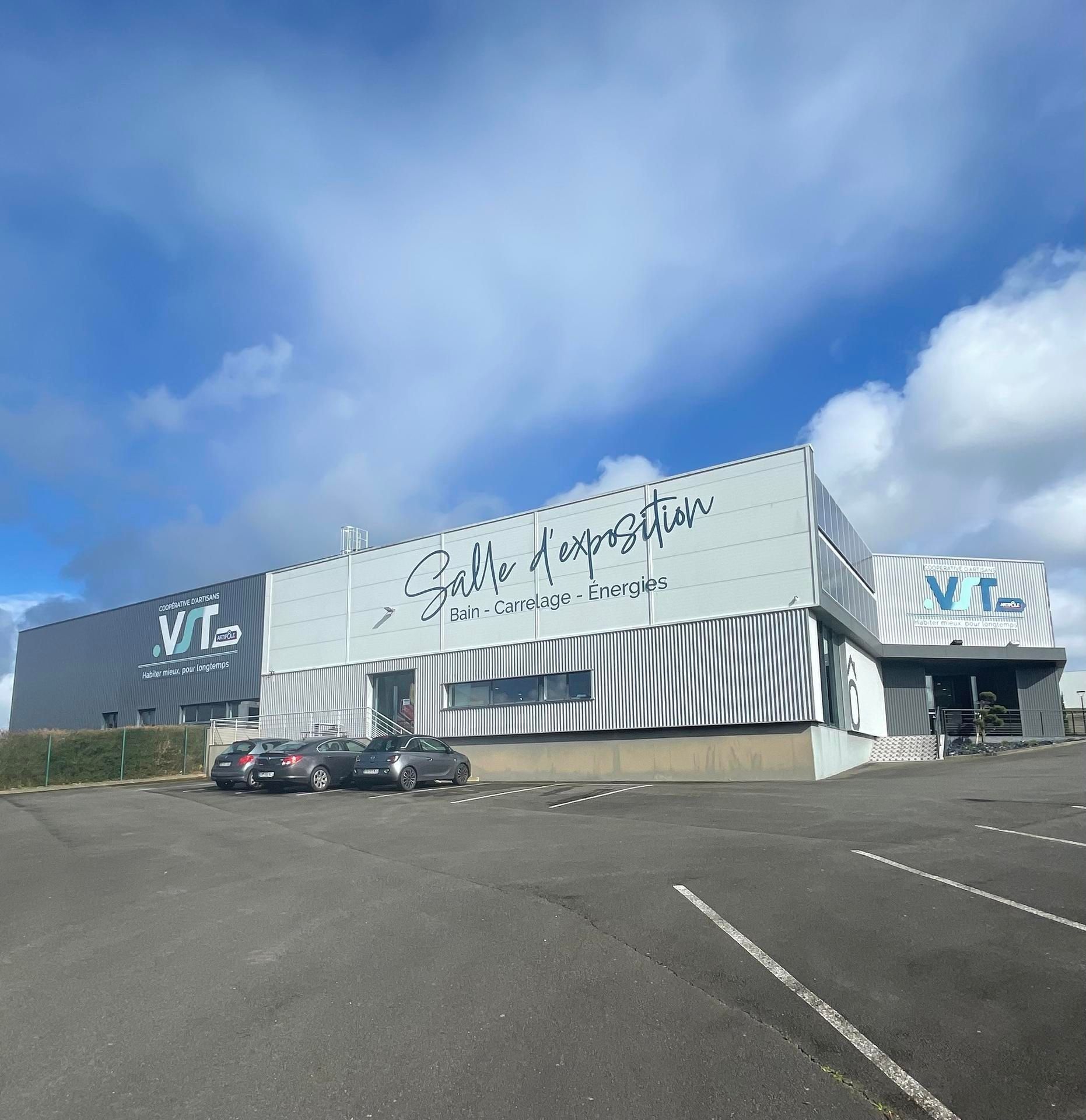 Un bâtiment commercial à la façade grise, un parking et une enseigne indiquant « Salle d'exposition » sous un ciel bleu nuageux.