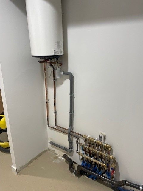Un réservoir de chauffe-eau blanc fixé au mur au-dessus de tuyaux de plomberie en cuivre et d'un système de collecteur avec des tubes bleus.