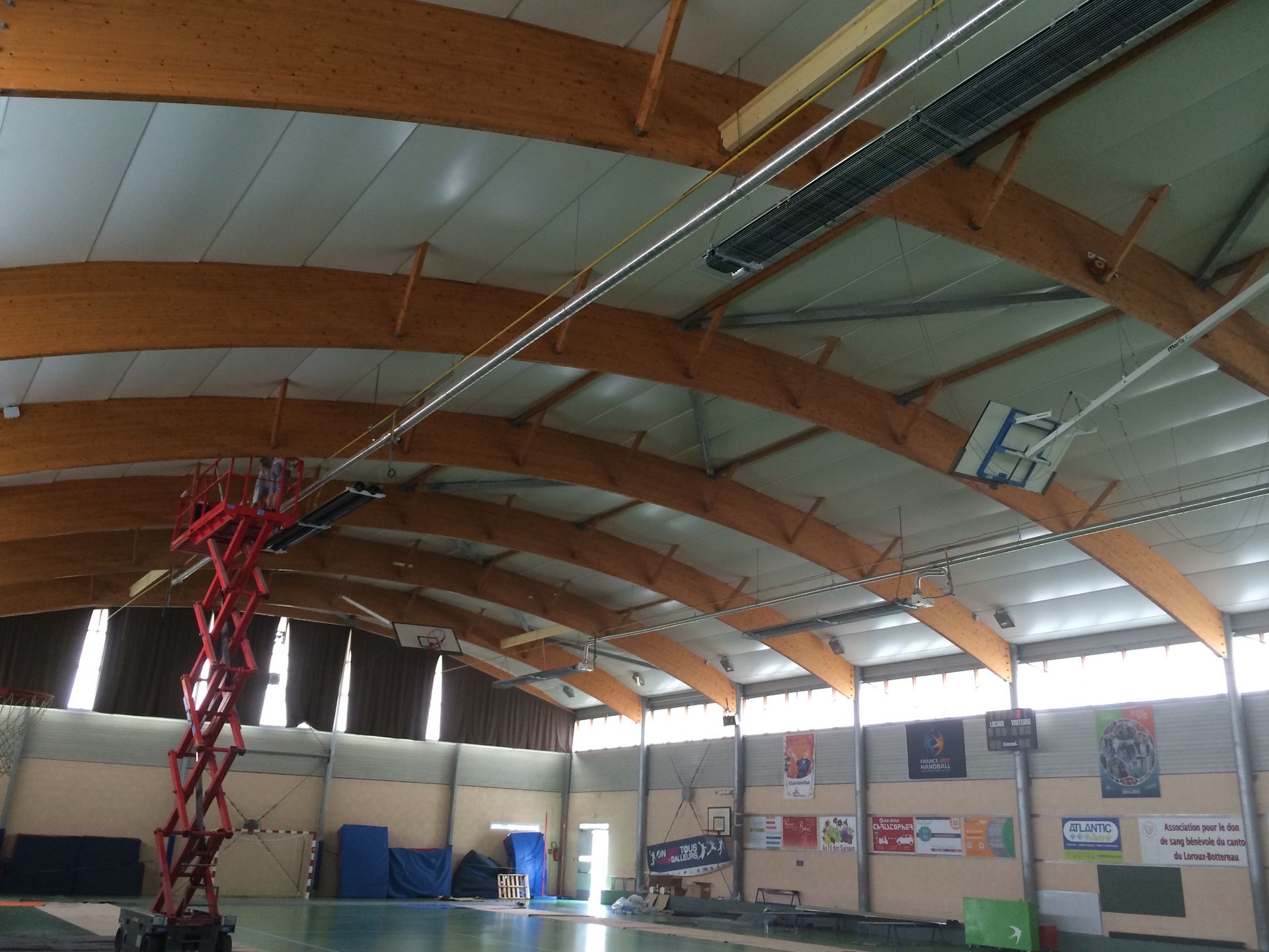 Dans un gymnase, une nacelle élévatrice rouge est déployée en hauteur pour atteindre les luminaires suspendus au plafond voûté à poutres en bois.