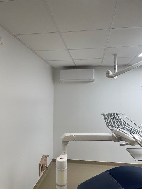 Une salle de soins dentaires avec un fauteuil bleu, un éclairage zénithal et une climatisation murale.