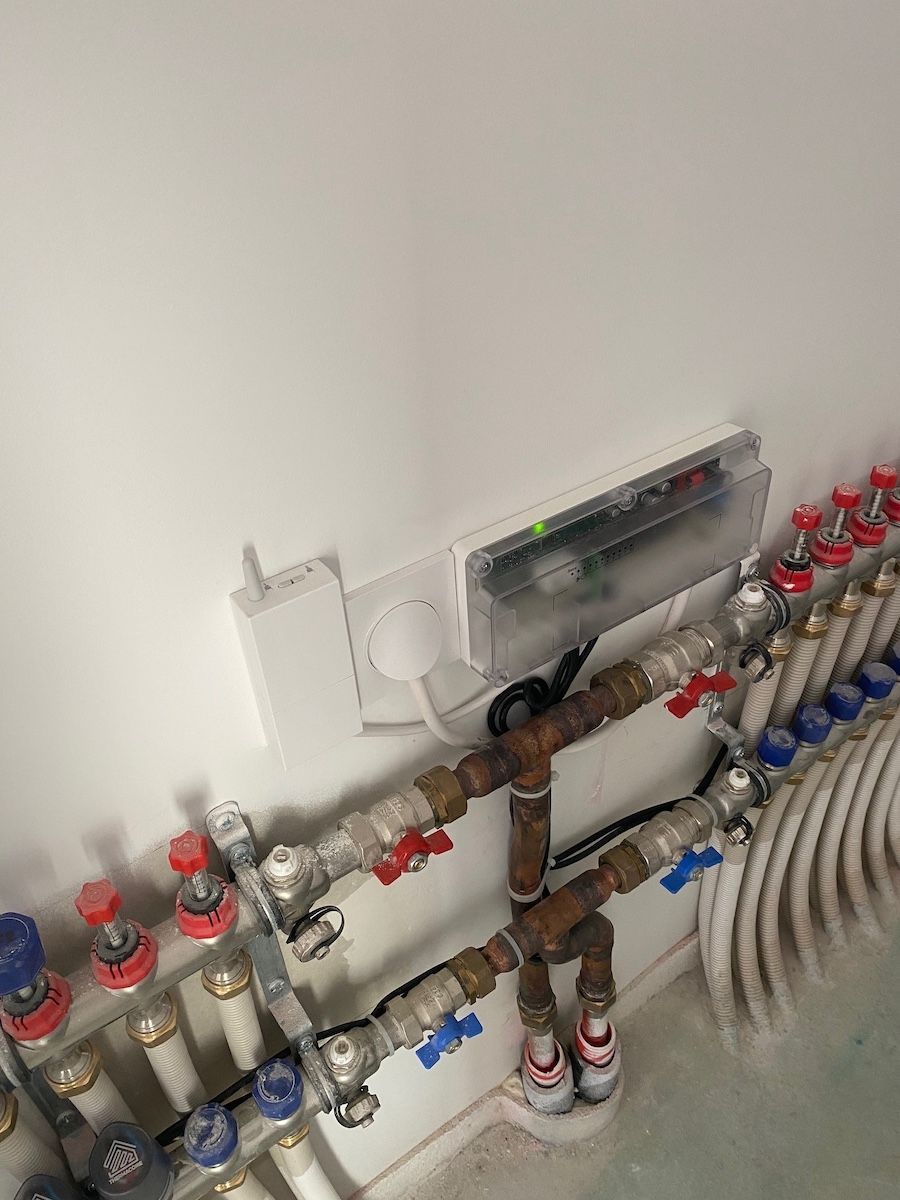 Un mur blanc sur lequel est installé un collecteur de chauffage au sol avec des tuyaux, des vannes et une unité de commande branchée sur une prise de courant.