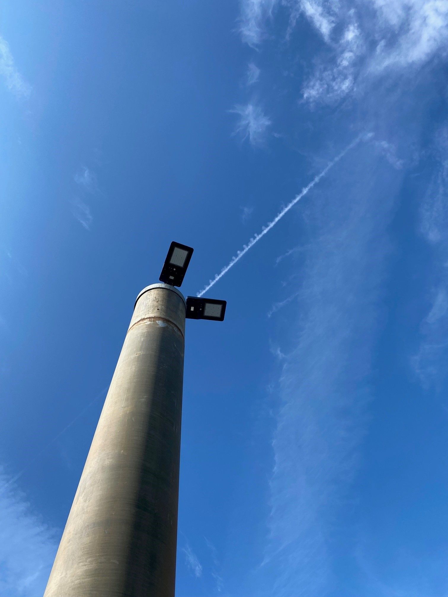 Un lampadaire en béton, équipé de deux luminaires LED, se dresse sur un ciel d'un bleu éclatant, traversé par une faible traînée de condensation d'avion.