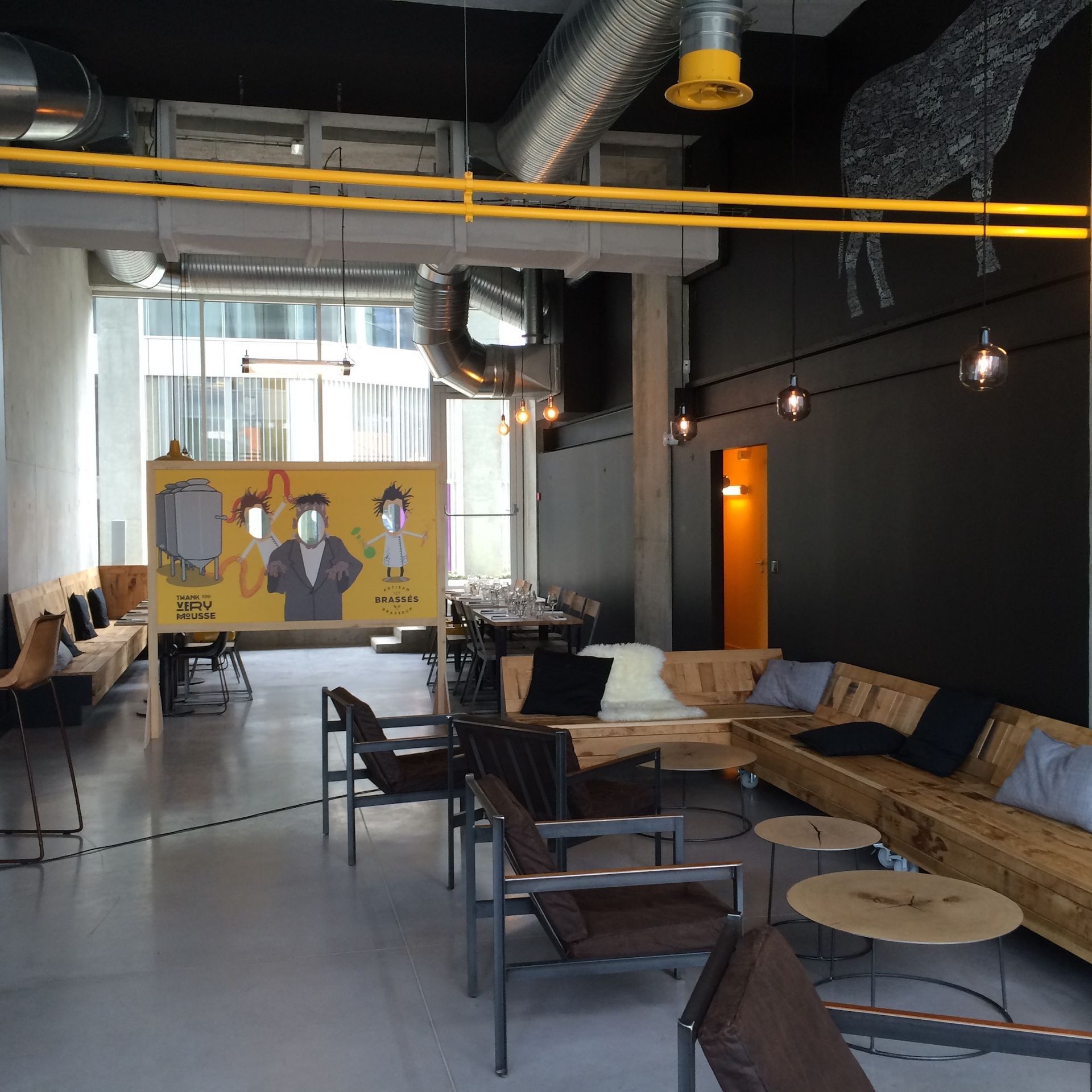 Un intérieur de style industriel avec des sièges lounge, des bancs en bois et une cloison jaune ornée d'une fresque murale dans un café.
