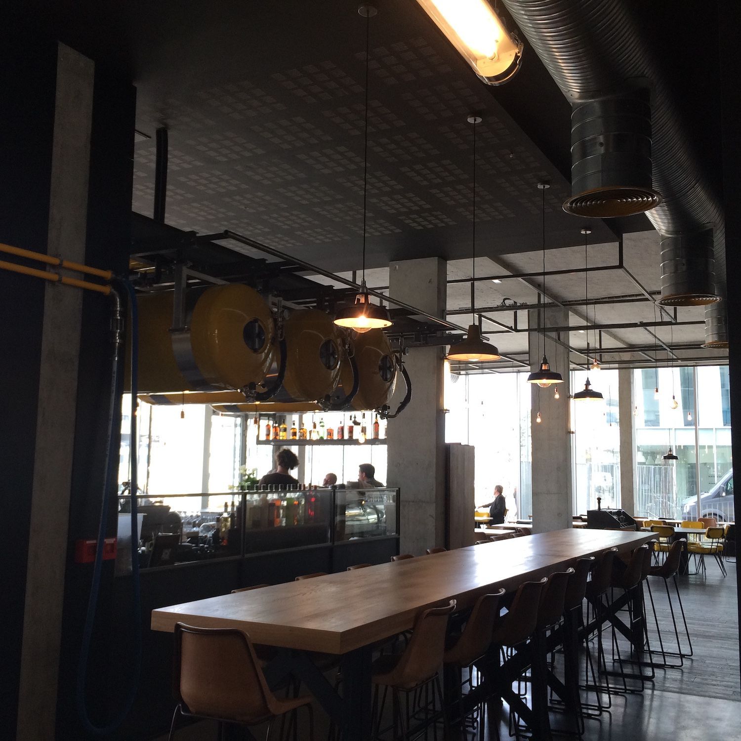 Une longue table en bois dans un restaurant à la décoration industrielle, avec des suspensions lumineuses et des fûts jaunes suspendus au plafond.