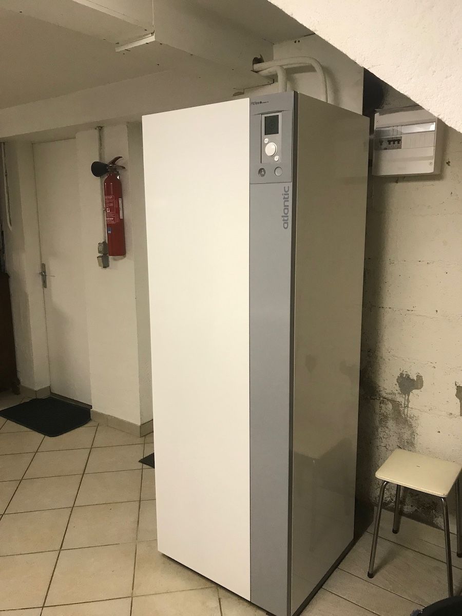 Une grande unité de pompe à chaleur grise et blanche se dresse sur un sol carrelé dans un sous-sol, près d'un extincteur et d'un disjoncteur.