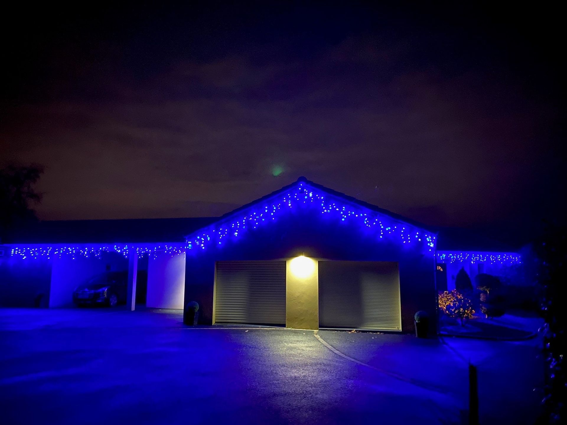 Vue extérieure sombre d'une maison la nuit, soulignée par des guirlandes lumineuses bleues le long du toit et du garage.