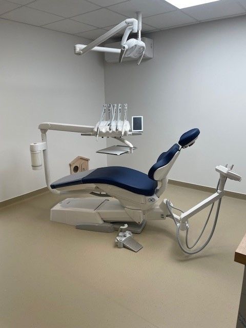 Un fauteuil d'examen dentaire avec un revêtement bleu, une lampe et des plateaux d'instruments dans une salle d'examen vide aux tons neutres.