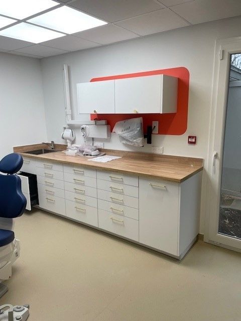 Cabinet dentaire avec armoires blanches, comptoir imitation bois, lavabo et fauteuil d'examen bleu sous éclairage encastré.