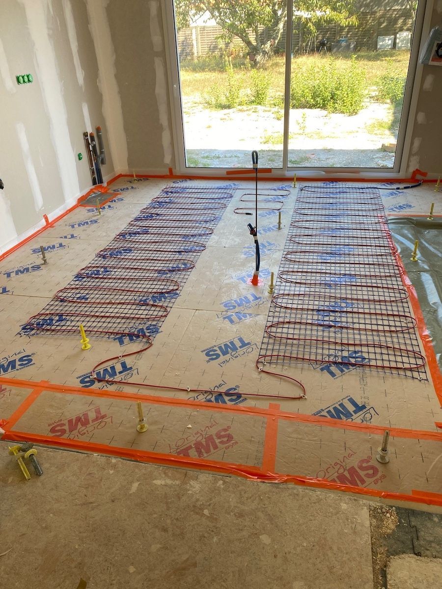 Des serpentins de chauffage au sol rayonnants sont installés sur des panneaux d'isolation sur un chantier de construction, avec une porte vitrée en arrière-plan.