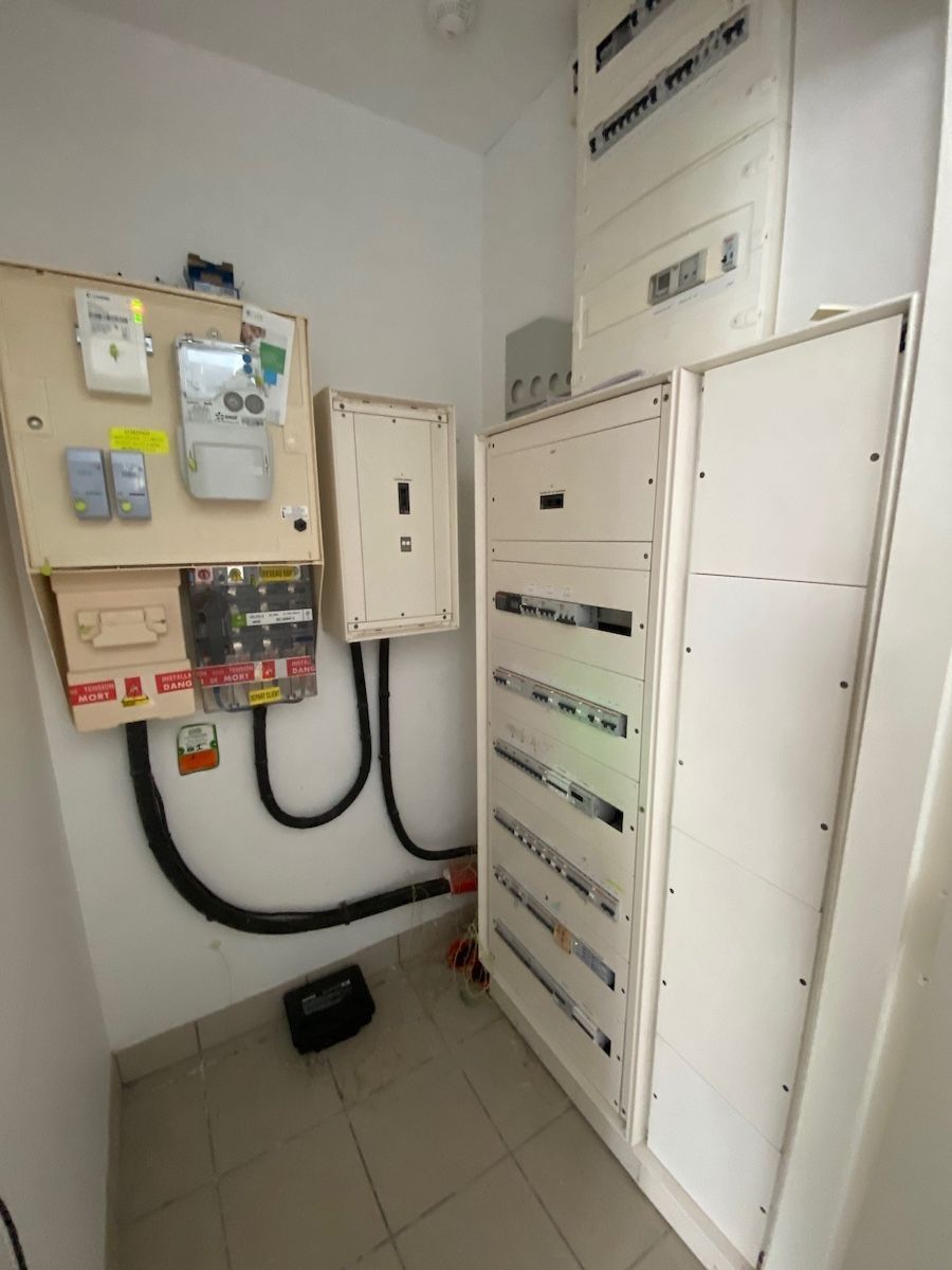 Local technique électrique avec plusieurs tableaux de disjoncteurs blancs muraux et un compteur électrique sur sol carrelé.