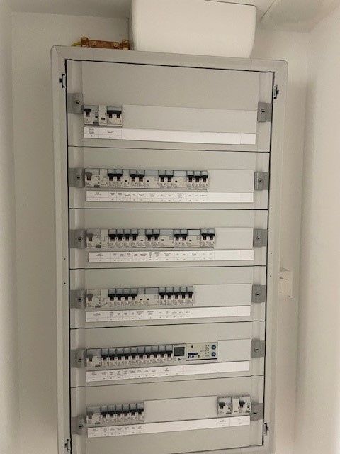 Un tableau électrique mural gris, comportant plusieurs rangées de disjoncteurs et d'interrupteurs.