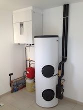 Un système de chauffage et d'eau chaude sanitaire blanc dans une pièce, comprenant une unité murale, un grand réservoir cylindrique et des tuyaux noirs.