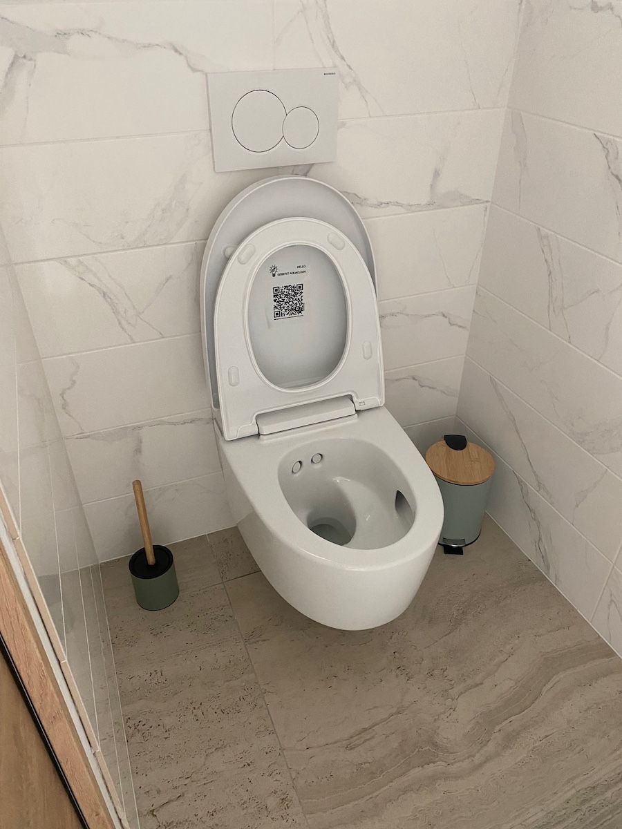 Des toilettes blanches suspendues avec un abattant ouvert, une brosse de toilette et une petite poubelle dans une salle de bains carrelée.
