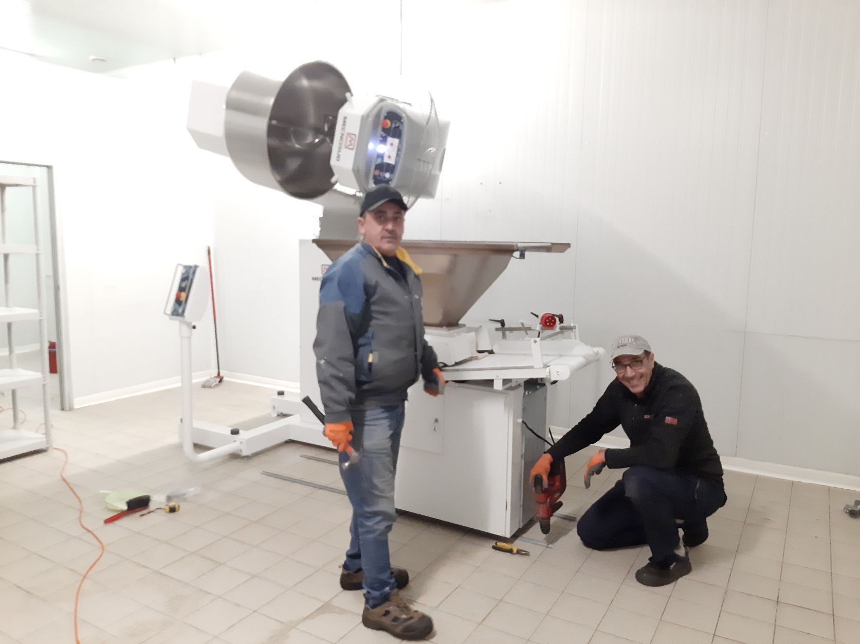 Deux hommes travaillent sur une machine industrielle blanche dans une pièce blanche. L'un est agenouillé, un outil à la main.
