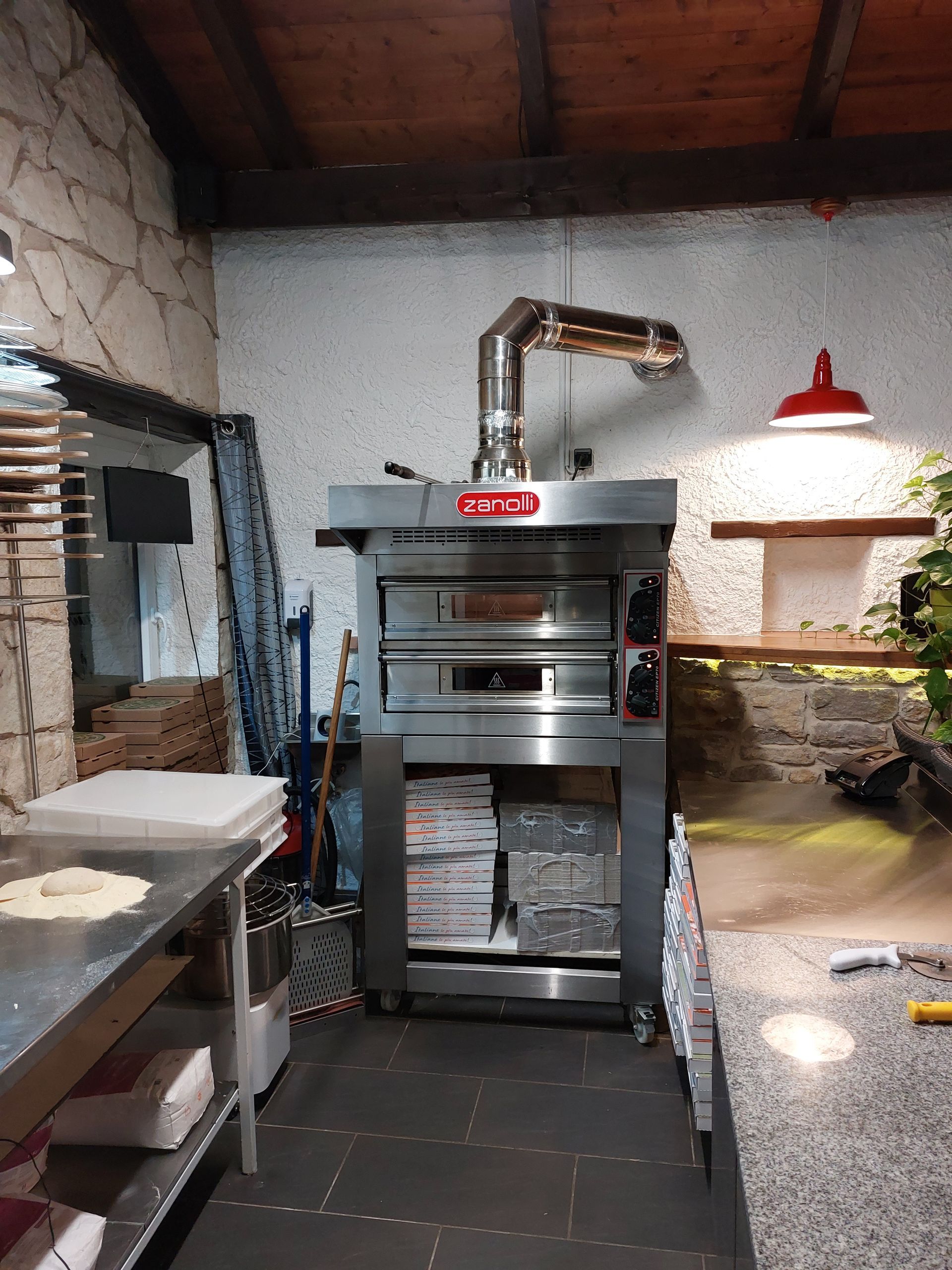 Four à pizza dans la cuisine ; acier inoxydable avec hotte aspirante. Murs en pierre et en brique, boîtes à pizza en dessous.