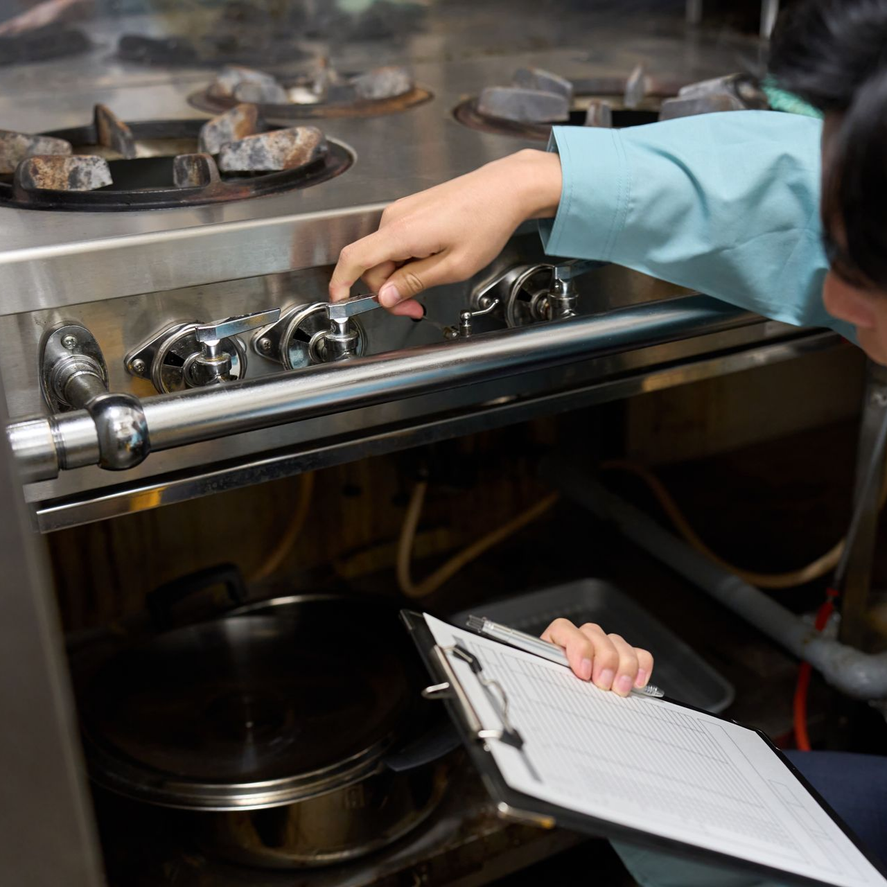 Personne portant une manche bleue inspectant une cuisinière de cuisine commerciale, tenant un presse-papiers.