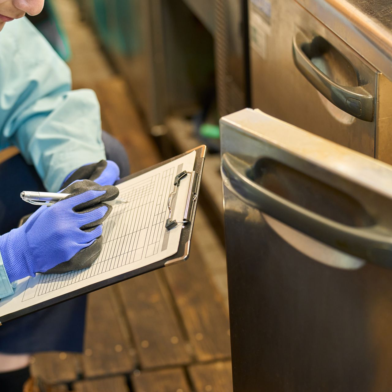 Une personne portant des gants bleus prend des notes sur un presse-papiers près d'une porte métallique.
