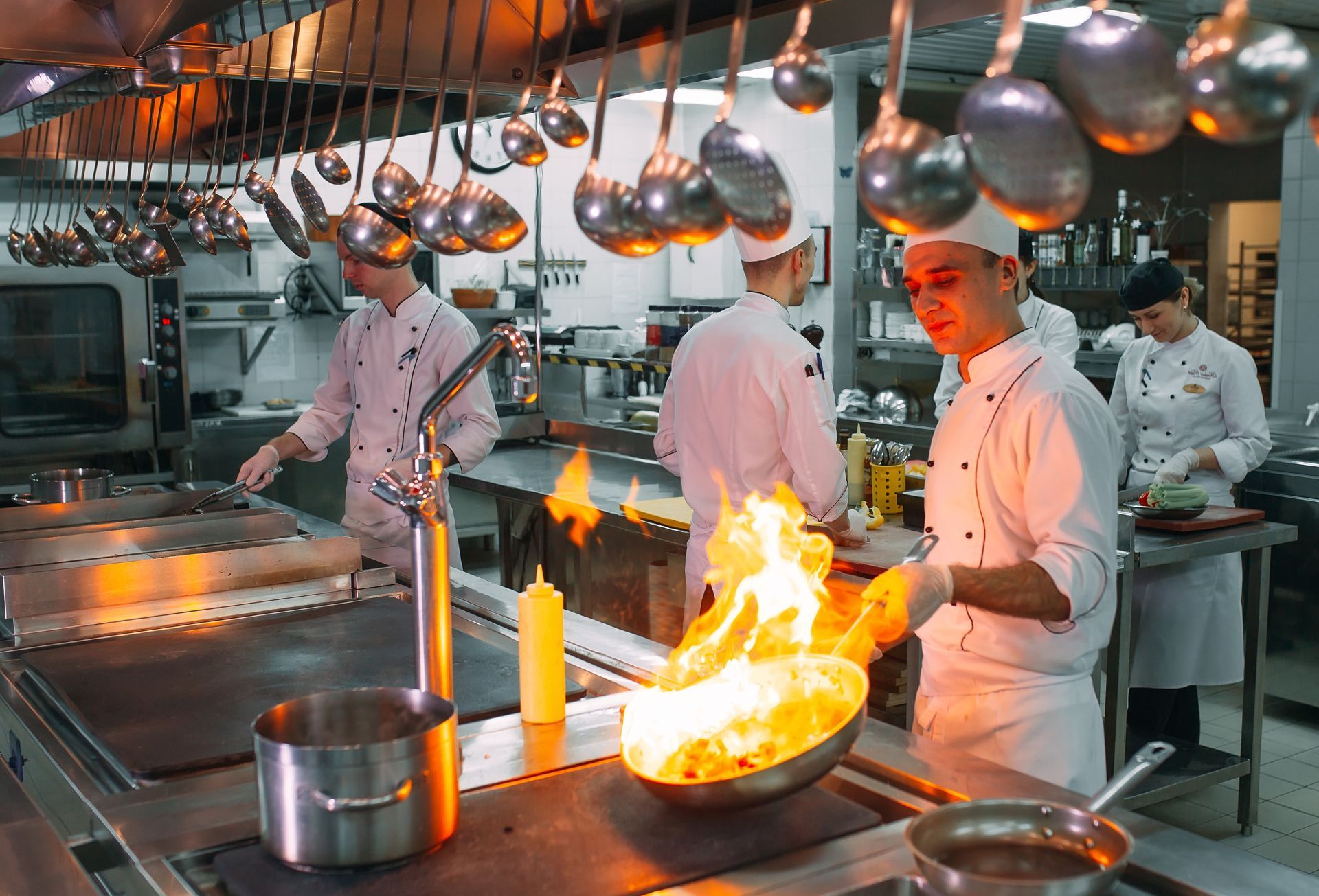 Des chefs dans une cuisine commerciale, l'un avec des flammes dans une poêle, d'autres aux fourneaux, des casseroles suspendues au-dessus.