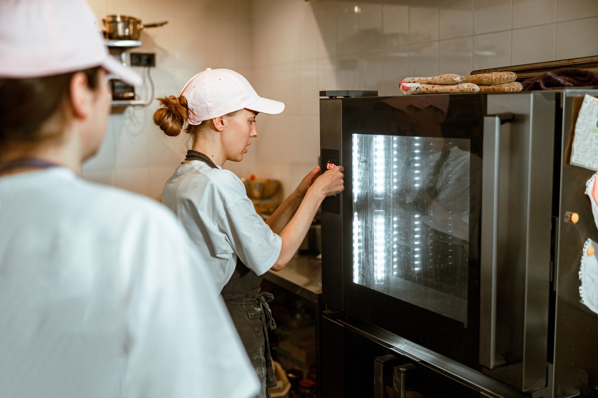 Deux boulangers dans une cuisine, l'un réglant un four. Tous deux portent des chemises blanches, des casquettes roses et des tabliers foncés.