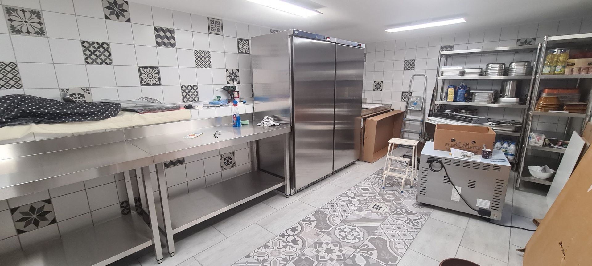 Une cuisine commerciale avec des tables en acier inoxydable, des réfrigérateurs, des étagères et des murs carrelés à motifs.