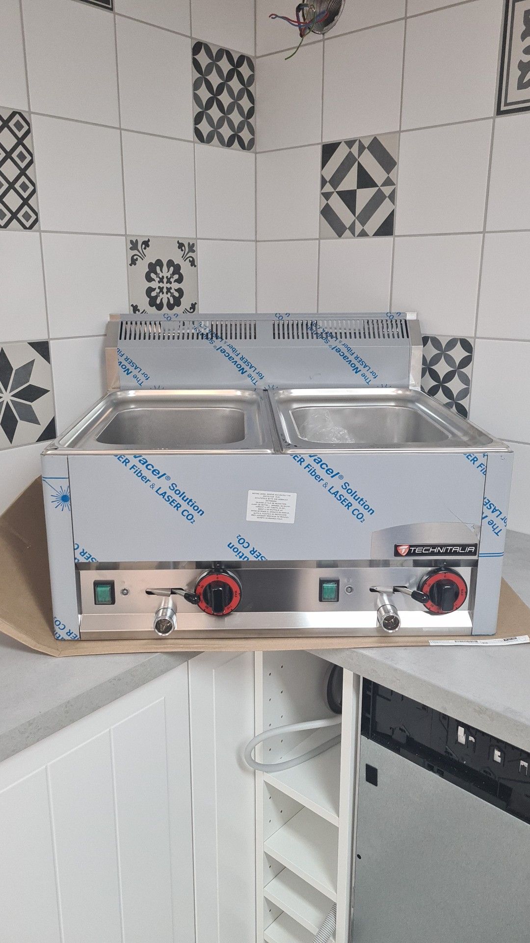 Meuble de cuisine commercial en acier inoxydable avec deux bacs chauffants, boutons rouges, contre un mur carrelé.