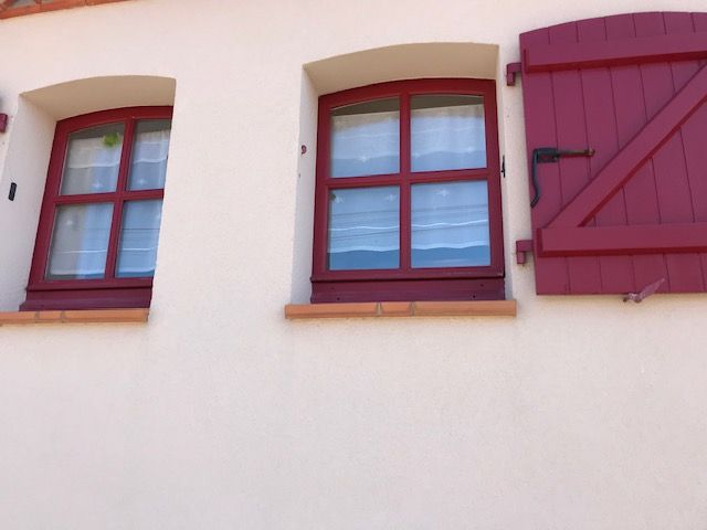 Deux fenêtres à cadre rouge et volets beiges sur un mur en stuc clair