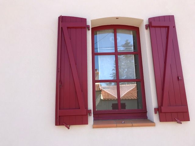 Fenêtre à volets rouges sur un mur en stuc blanc, avec reflets d'arbres et de toits.