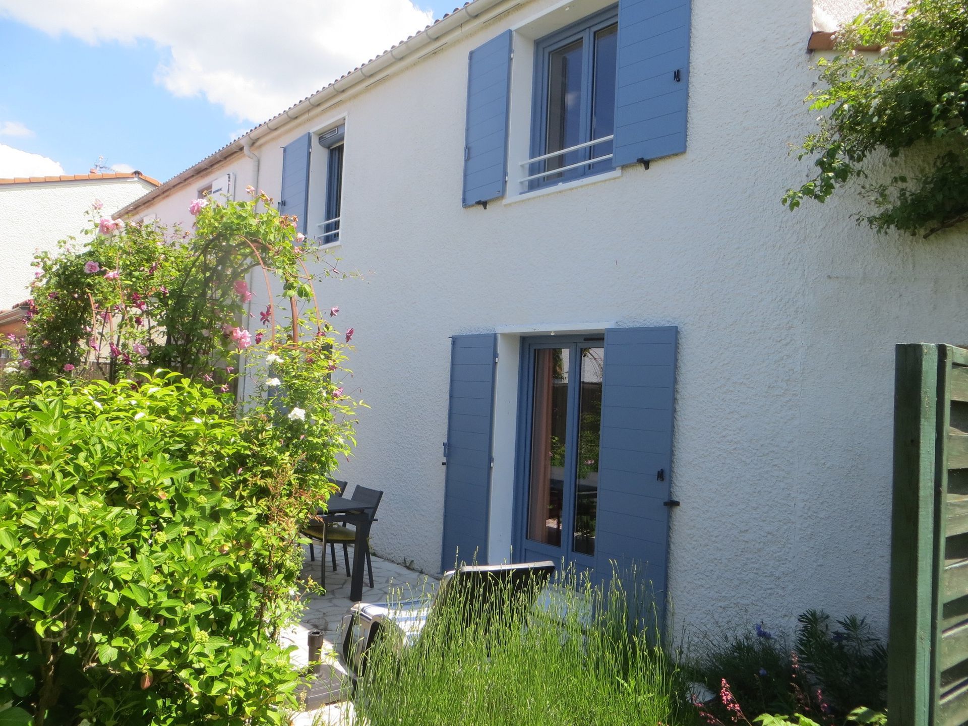 Maison en stuc blanc avec volets bleus et patio, partiellement dissimulée par la verdure et les fleurs du jardin.