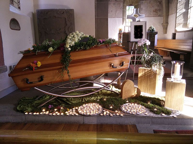 Holzsarg mit weißem Blumenschmuck erhöht aufgebahrt in einer Kapelle