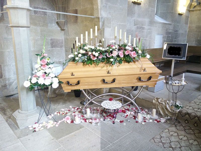 heller Sarg mit Blumenschmuck in Kapelle erhöht aufgebahrt mit Kerzen auf dem Boden und Rosenblättern