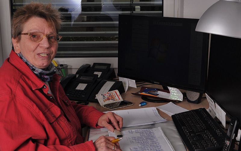 Eine Frau in einer roten Jacke sitzt an einem Schreibtisch vor einem Computer.