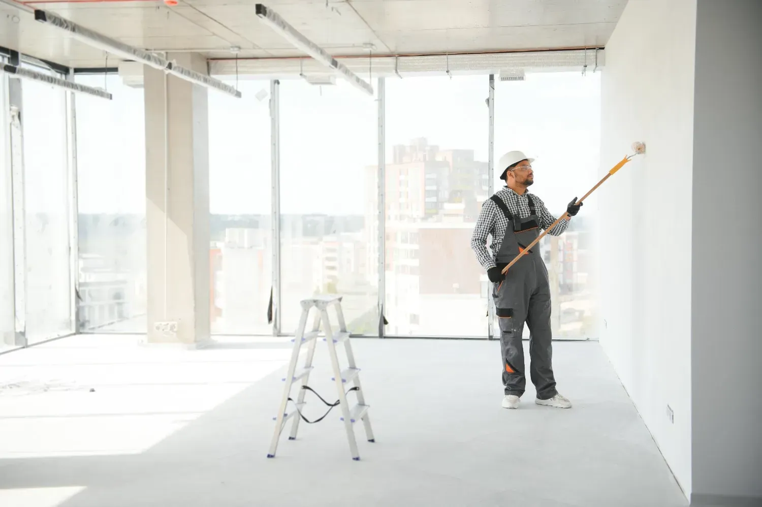 Pintor utilizando un rodillo en una habitación luminosa y vacía con una escalera y grandes ventanas.