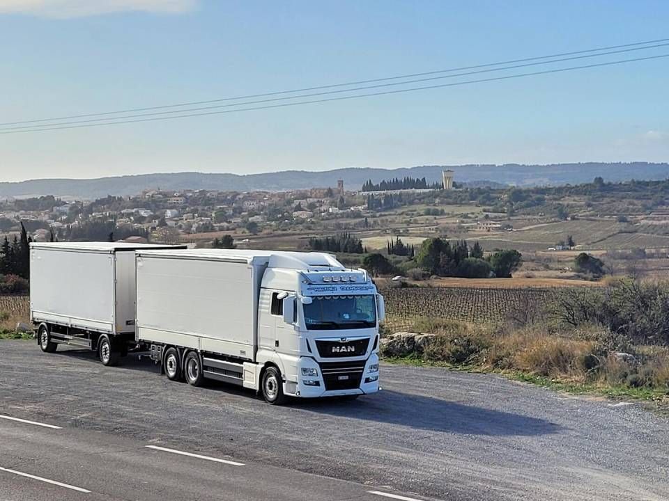 Weisser Sattelzug mit zwei Anhängern parkt auf einer Strasse, ländliche Landschaft mit einer Stadt im Hintergrund.