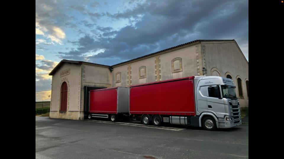 Ein silberner Sattelzug mit roten Anhängern parkt vor einem hellen Gebäude unter bewölktem Himmel.