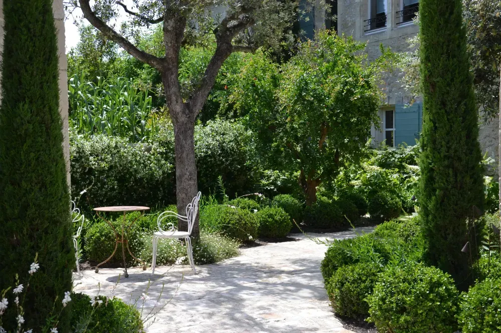 Allée ensoleillée dans un jardin luxuriant, avec un chemin de pierres et une petite table blanche et des chaises près d’un bâtiment