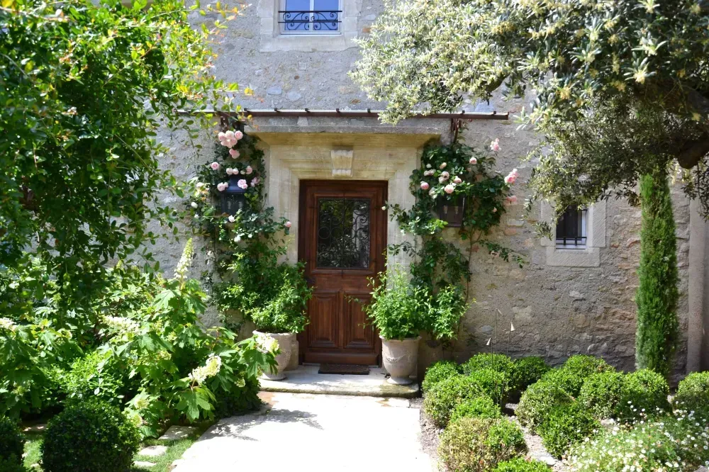 Entrée d’une maison en pierre avec une porte en bois, entourée d’un jardin verdoyant et d’arbres